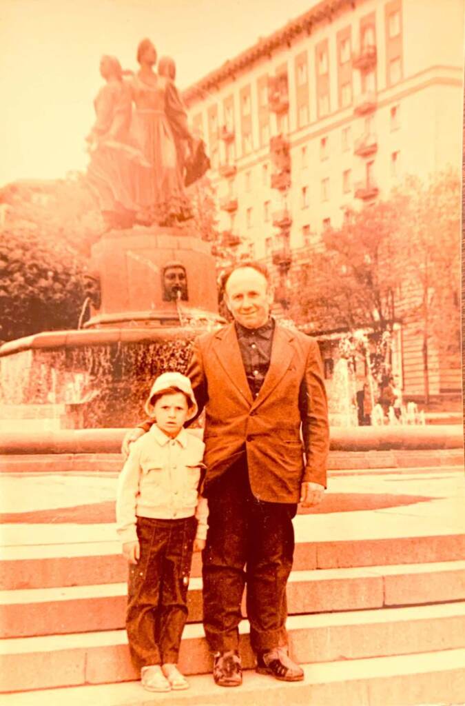 Doc-Mar-29-2022-9-59-PM-712×1080 A sepia-toned photograph shows an older man standing with a young boy in front of a fountain.