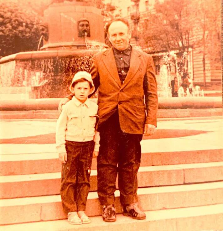 A sepia-toned photograph shows an older man standing with a young boy in front of a fountain.
