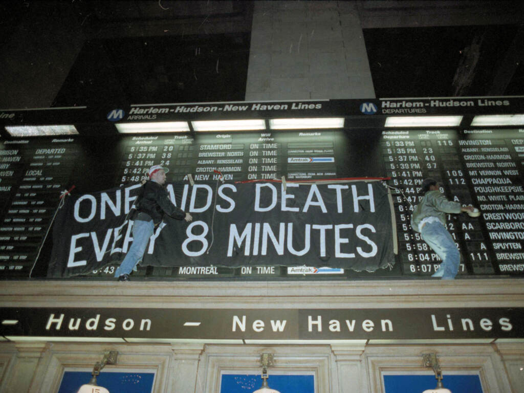There are parallels between COVID and HIV. Despite effective treatment and prevention tools, preventable deaths continue because of difficulties reaching out to and educating people about the tools. And even as the country seems determined to move on from the pandemic, as of April 2022, someone dies of COVD-19 every four minutes in the U.S. Members of the group Act-Up hang a banner across the train schedule board in New York's Grand Central Terminal