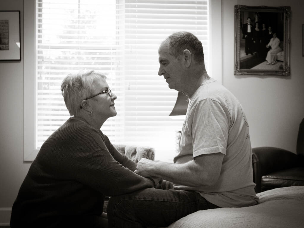 Mary Daniel took a dishwasher job at her husband’s Florida memory care facility to see him during the initial coronavirus lockdown. She has been fighting for visitation rights ever since. Mary Daniel kneels down by her husband, seated on a bed at his Florida memory care facility.