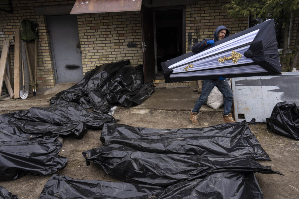 A man carries a coffin next to plastic bags with corpses of civilians
