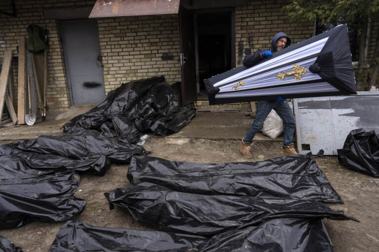 A man carries a coffin next to plastic bags with corpses of civilians