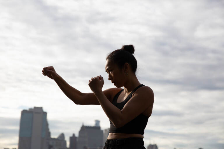 Jess Ng is shown in a defensive stance, with city buildings and sky behind her.