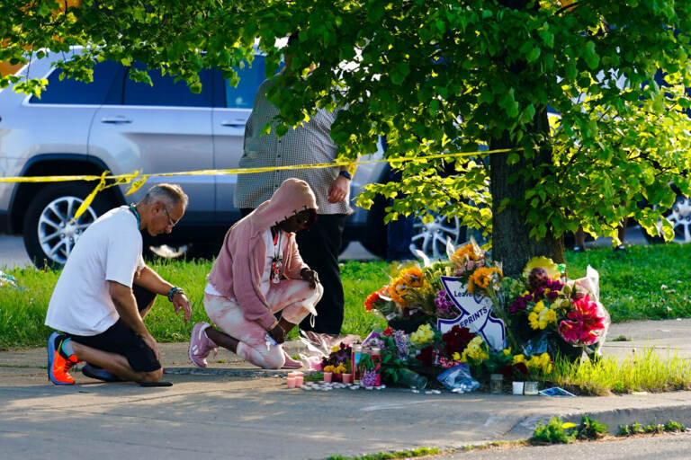 Two people kneel on the ground in front of a memorial made of flowers.