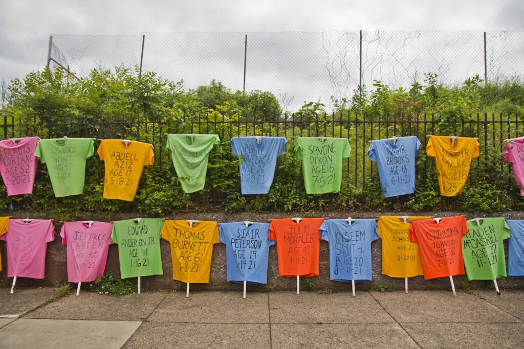 day of serenity k paynter 2022 05 15 (1 of 1)-8 (1) Rows of T-shirts bearing the name of Philadelphians lost to gun violence are on display with a fence and bushes in the background.