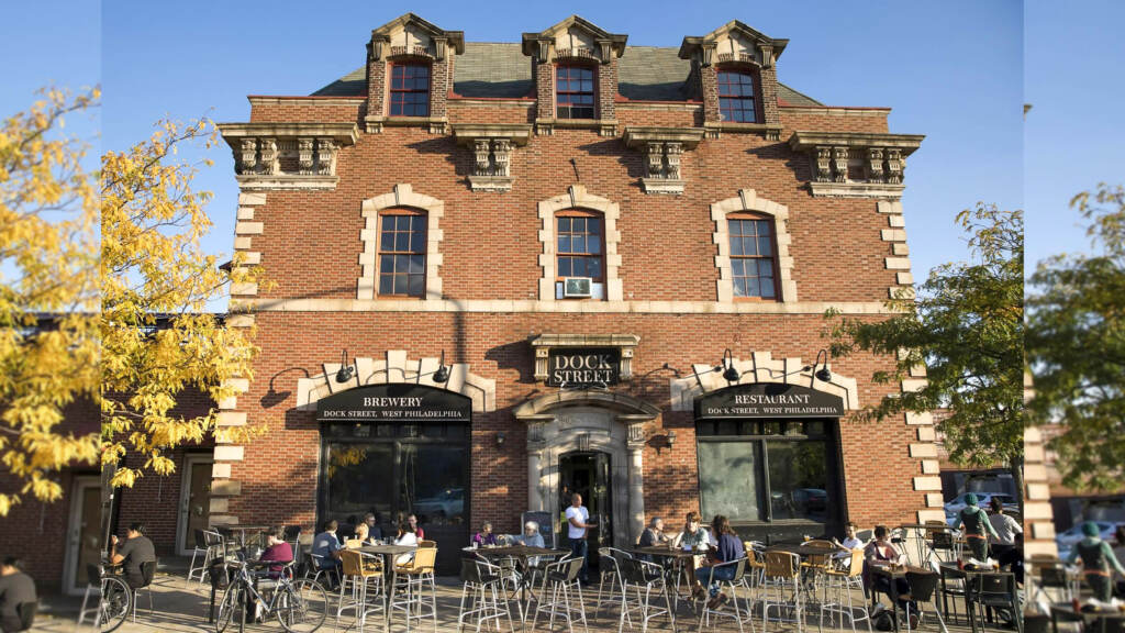 dockstreetbrewing-westphilly The brick building which currently houses Dock Street Brewing in West Philly is visible in the early evening sunlight. People are sitting at tables outside.