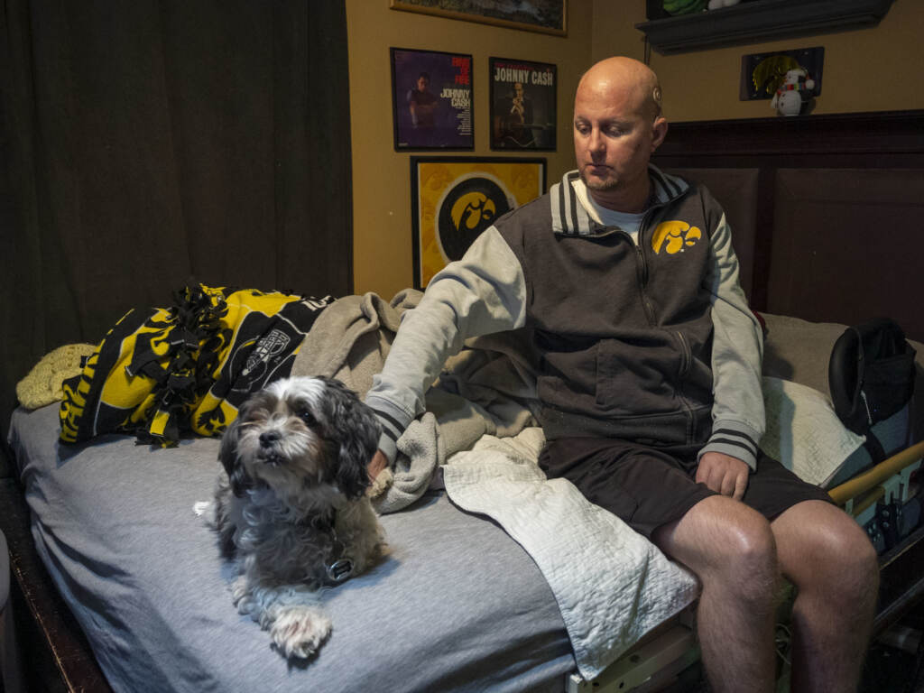 Jon Miller sits in his bedroom with his dog, Carlos, whom he received as a present for successfully completing cancer treatment a decade ago. Miller sustained severe brain damage, and requires the help of home health aides to continue living in his home. Jon Miller sits in his bedroom with his dog, Carlos, whom he received as a present for successfully completing cancer treatment a decade ago.
