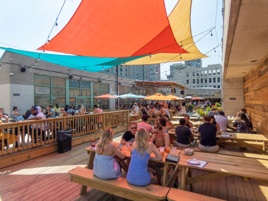 libertypoint-waterfront-10 People sit at wooden picnic tables under a canopy protecting them from the sun.