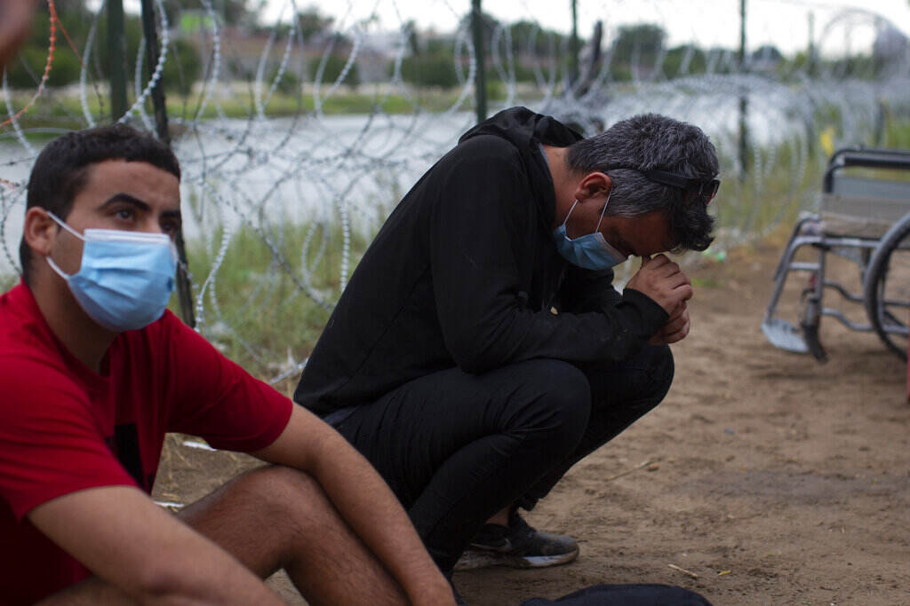 Border Asylum Limits A man looks up with a face mask on. Another man squats close to the ground beside him, his face in his hands.