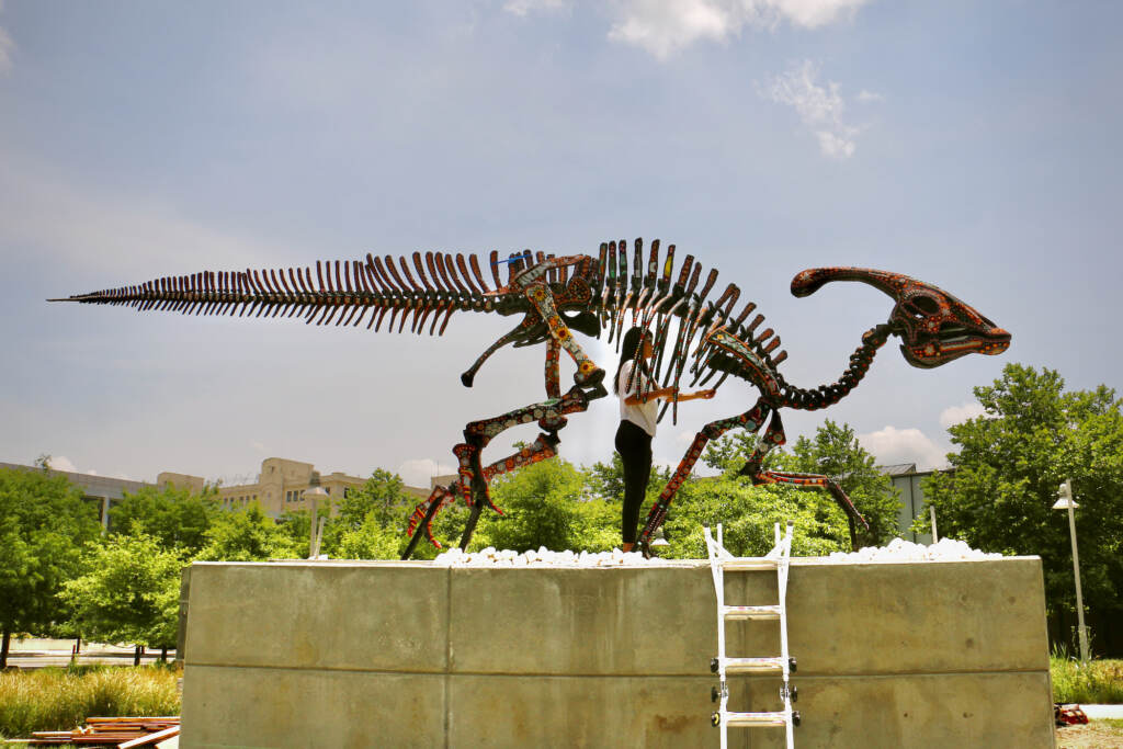 2022 06 08-e lee-marianela fuentes-philadelphia navy yard-parasauralophus art ribcage Artist Marianela Fuentes stands in the ribcage of her sculpture, ''Alpha Sacred Beings (The Order of Creation),'' mounted on a six foot concrete plinth in Philadelphia Navy Yard. (Emma Lee/WHYY)