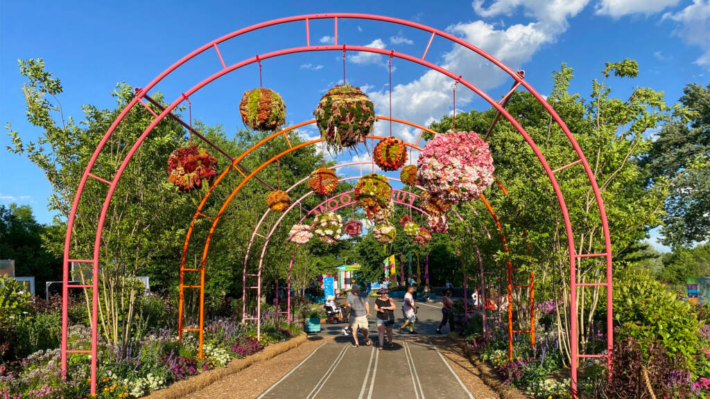 flowershow2022-fdrpark-12crop-1 An arching trellis with flowers is shown over a walkway with trees and a blue sky in the background.