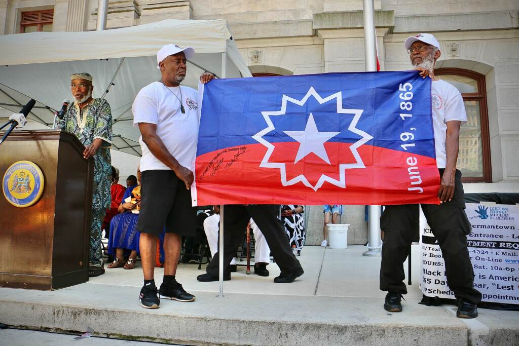 juneteenth-flag-raising-e-lee-20220606-6 Members of the Black Male Community Council hold up the official flag of Juneteenth while Ben Haith (left) the creator of that flag describes what inspired him. Haith was the guest of honor at the flag raising ceremony at Philadelphia City Hall. (Emma Lee/WHYY)