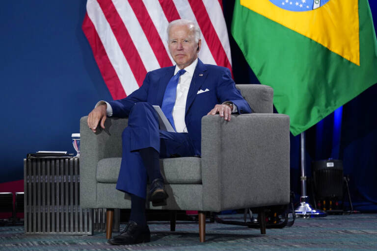 President Biden sits in a chair, with the U.S. and Brazilian flags behind him, at the Summit of the Americas.