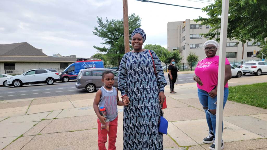 summer-enrichment-classes-west-philadelphia A mother stands holding her child's hand and smiling, with the sidewalk and street in the background.