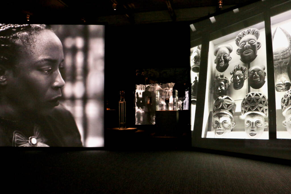 A video panel of a woman looking off to the side is next to another video panel showing an African art collection.