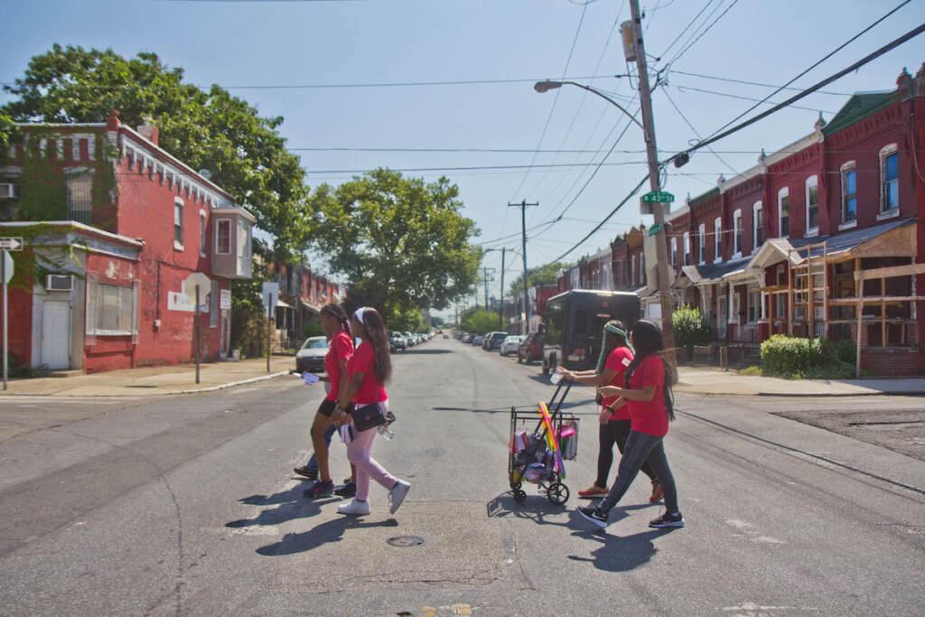 2022 07 11 k paynter play streets (1 of 1)-6 People cross a street in West Philadelphia, with brick rowhomes lining either side of the street.