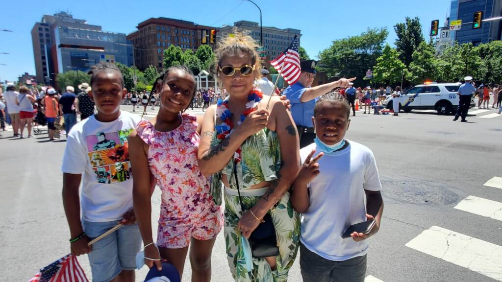 A group of people stand on the side of the street, holding the American flag and smiling.