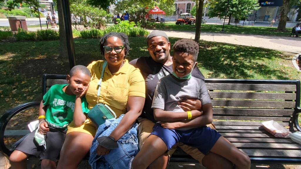 A smiling family, two parents and their two sons, sit on a bench in the shade.