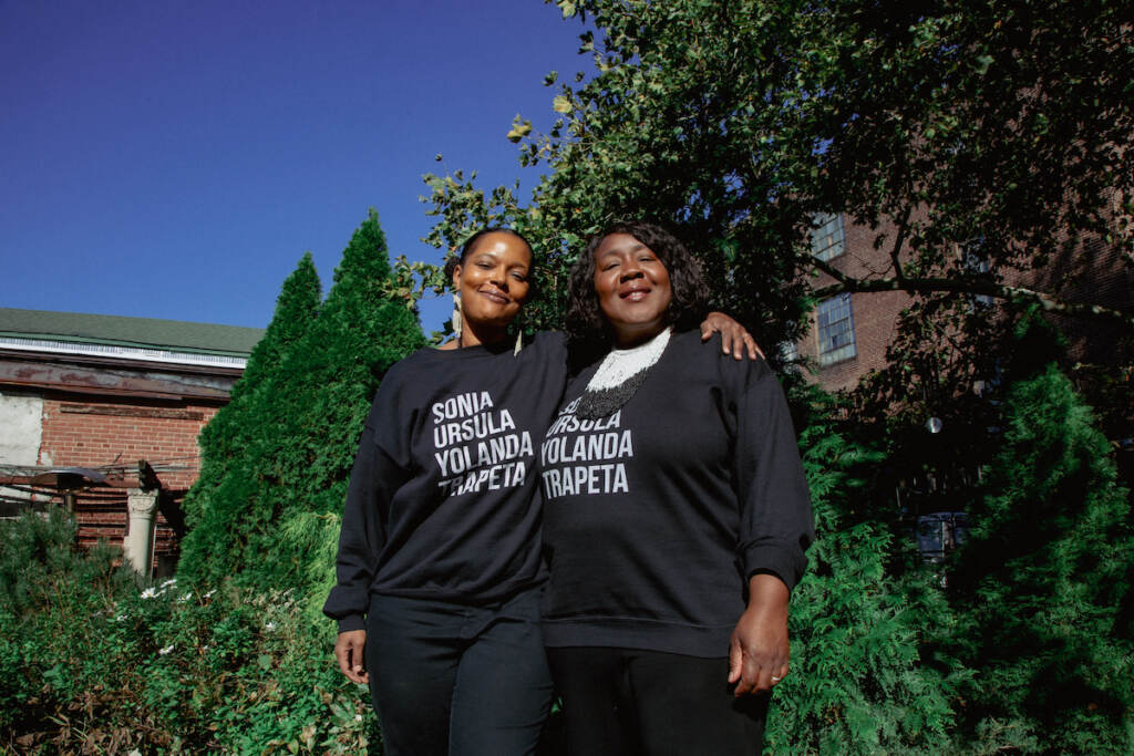 Regeneration_ConsenSIS_Headshots00009(1) Two women stand together, facing the camera and smiling, with trees and a brick building visible in the background.