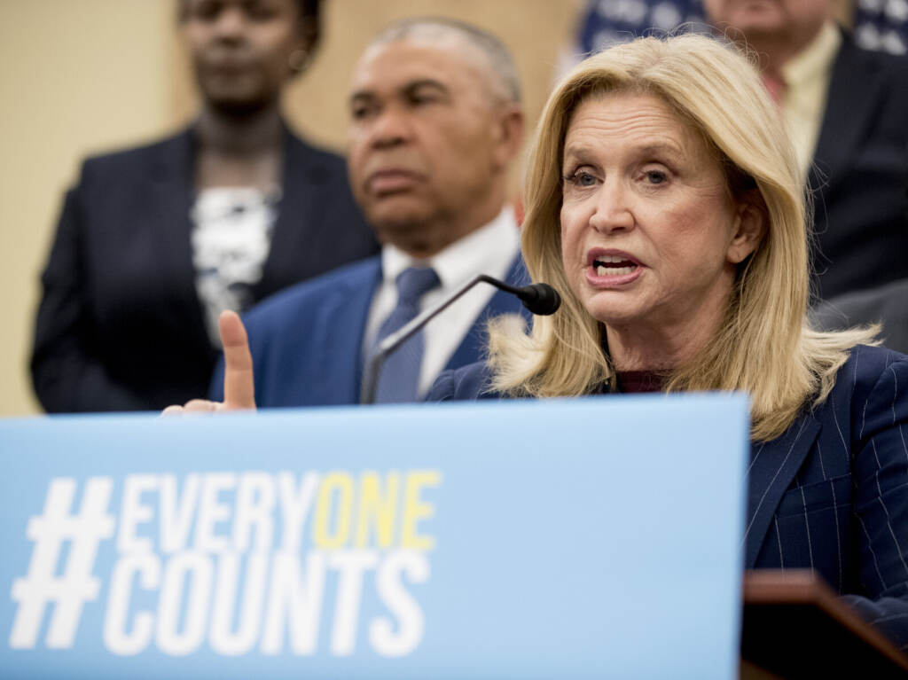 Carolyn Maloney Someone speaks at a podium with a sign reading,