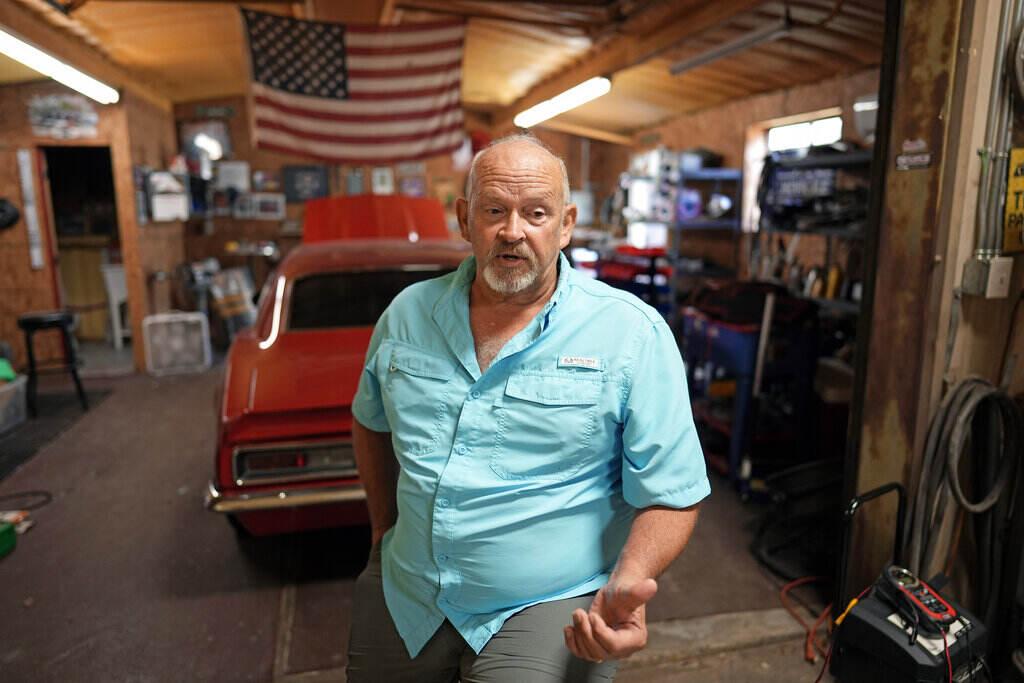 A man stands, speaking, in the foreground of the photo, with a red car and the American flag visible behind him.