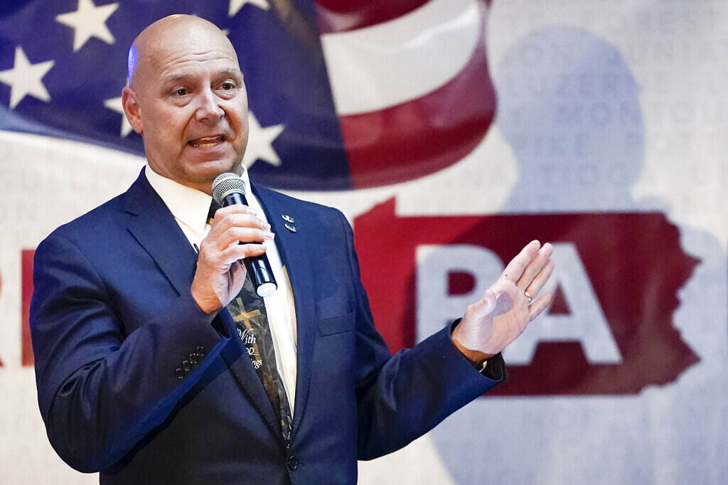 Doug Mastriano speaks in front of an image of the American flag and PA written in the background.