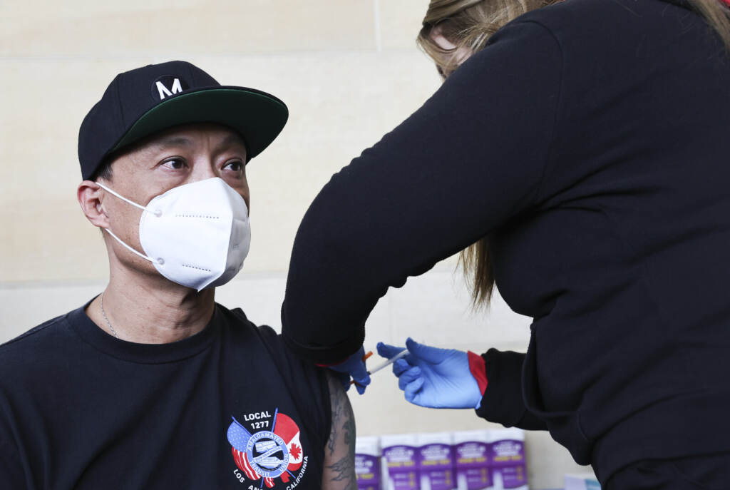 A person wearing a face masks looks forward as a healthcare professional gives them a vaccine in their arm.