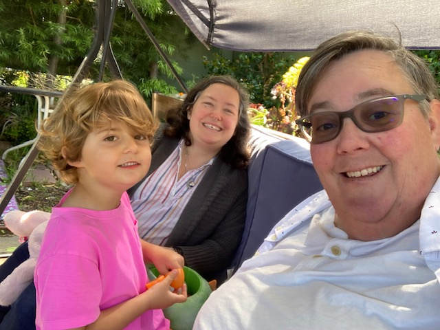 A selfie of Beth Kenny (foreground), their wife Adina (middle), and their child Vyla sitting in their backyard in Alameda, Calif. Since the lifting of COVID safety measures, Kenny and their family have had to pull back from indoor activities, and they struggle to explain why to Vyla. A selfie of Beth Kenny (foreground), their wife Adina (middle), and their child Vyla sitting in their backyard