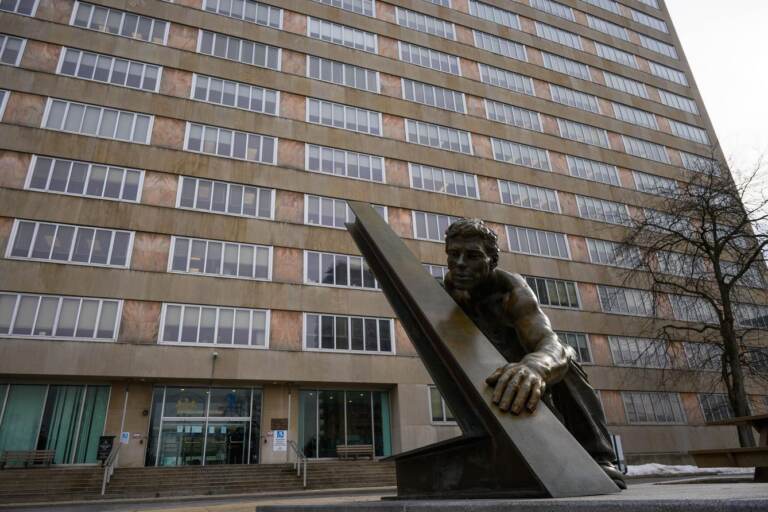 A statue of someone holding up something is visible in front of a tall building.