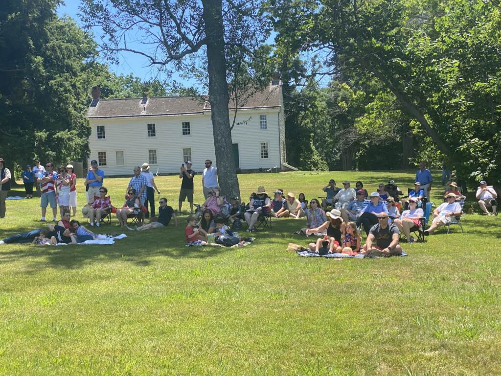 princeton-battlefield-declaration-independence A crowd sits on green grass, with some trees and a building in the background.