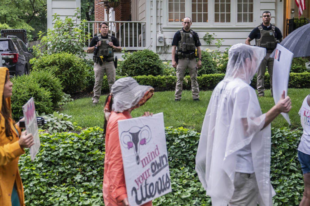 Abortion Rights Activists Protest Outside Justices’ Houses People hold signs and march, with officers standing in the background between protesters and a house.