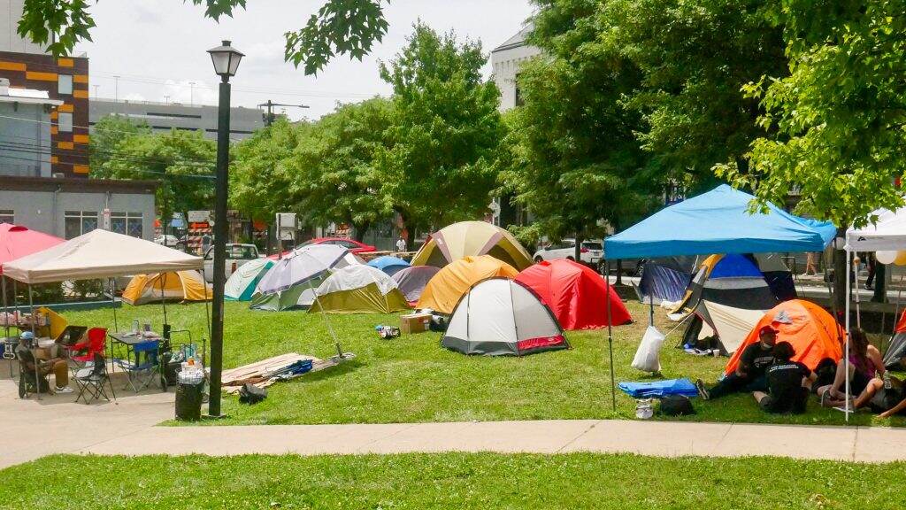 westphilly-uctownhomes-encampment-01-1024×576 Tents are pitched on green grass. A sidwalk is visible in the foreground and a building and trees are visible in the background.