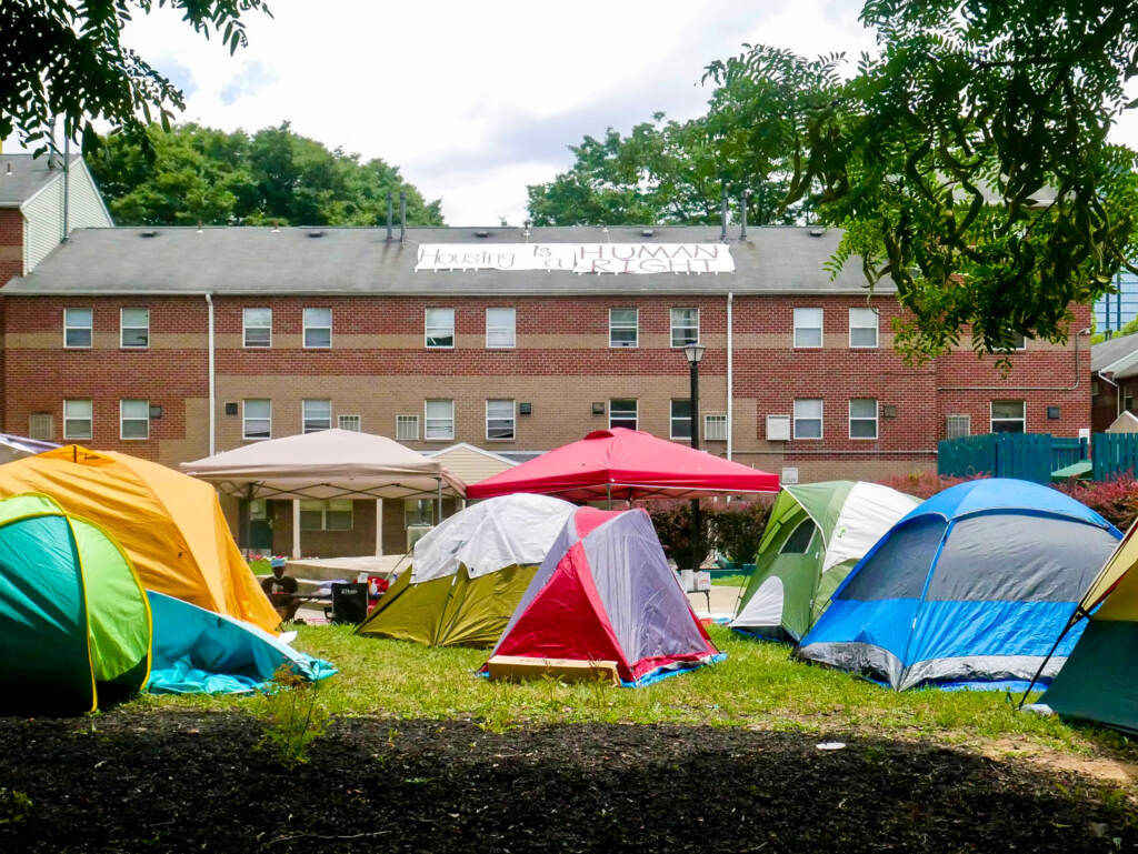 westphilly-uctownhomes-encampment-02 Tents are visible on a lawn in front of a red-brick building.