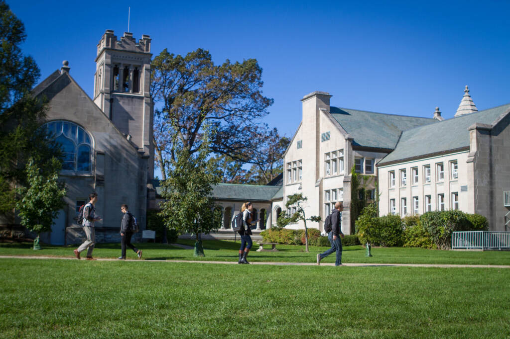 colleges-prepare-monkeypox Students walk across grass on a green campus with buildings in the background.