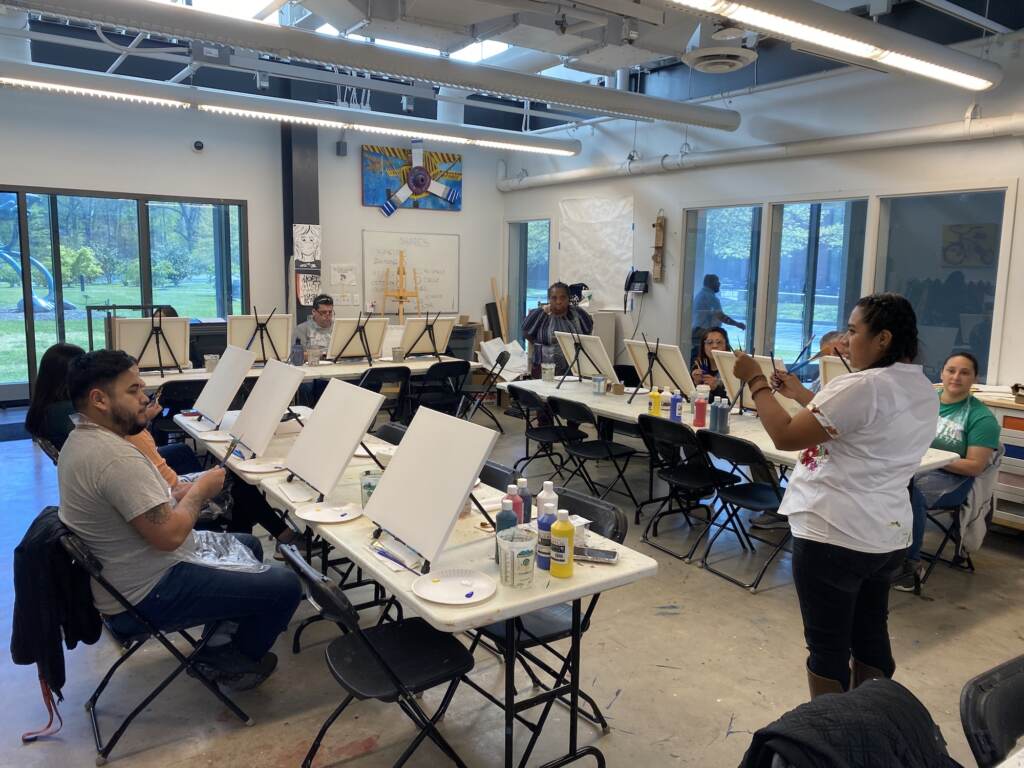 delaware-art-museum-healing-arts-1 A person speaks and gestures at the front of a room, where students are sitting before blank easels.