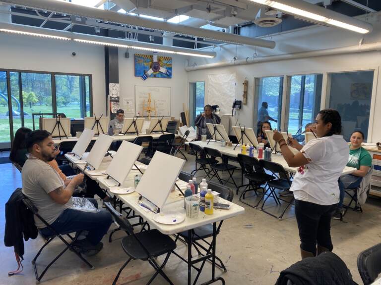 A person speaks and gestures at the front of a room, where students are sitting before blank easels.