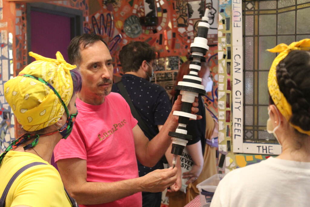magic-gardens-recycle-art-event-1 The artist, in a pink T-shirt in the center, holds up plastic recycled art while two people, backs to the camera, listen.