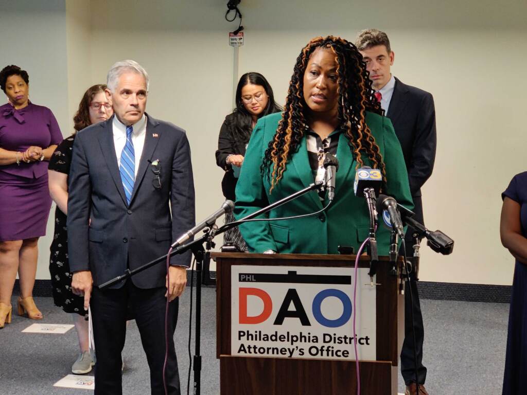 network-fight-elder-abuse A woman in a green blouse speaks at a podium with a sign in front which reads DAO. People stand in the background.