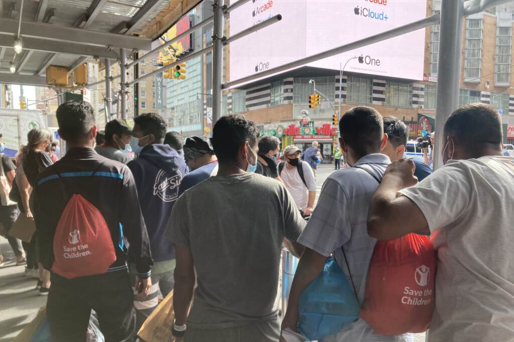 Asylum-seekers who have recently arrived in New York on a bus from Texas wait for ride-share vehicles, organized by a mutual aid group, to take them into the city. A group of migrants arriving in New York City are shown from behind as they look out on a city street.