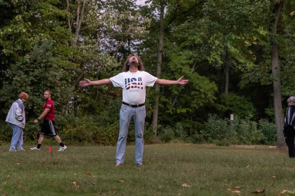 A man stands in the middle of a green field, trees behind him, looking up towards the sky with arms outstretched.