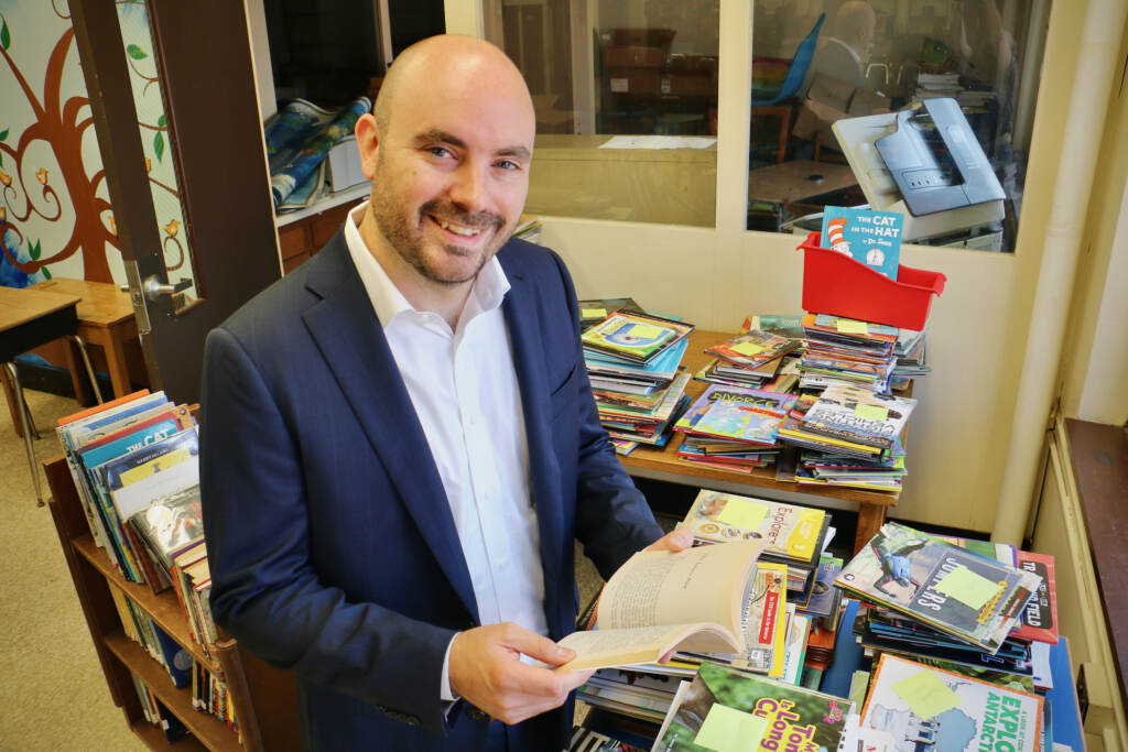 A man holds an open book in a classroom surrounded by stacks of books.