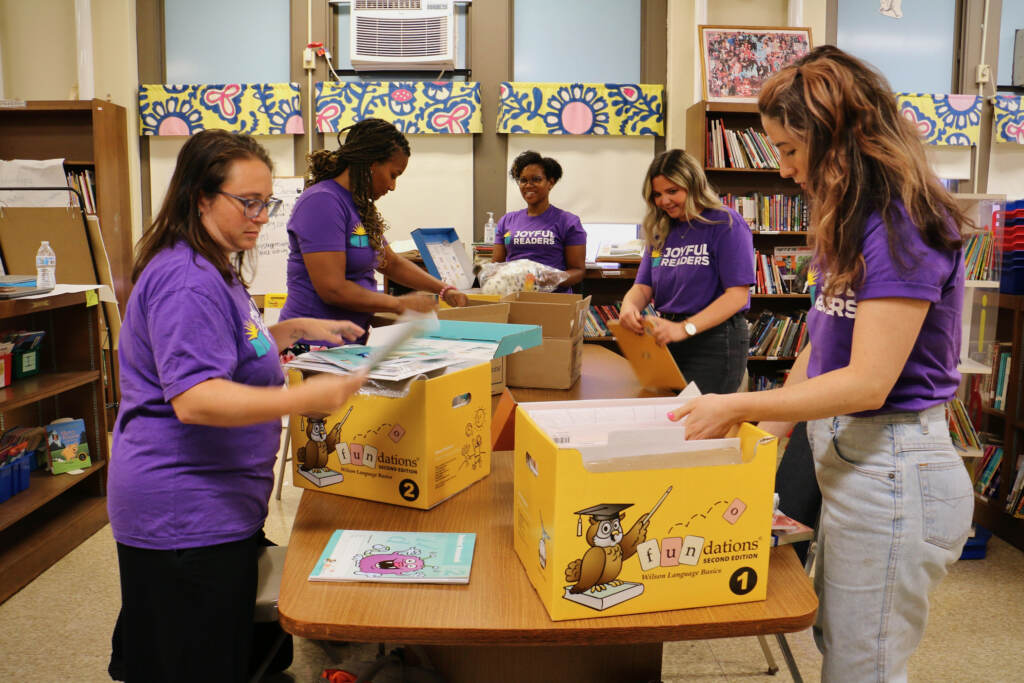 A group of people in purple shirts gather around a long table with boxes full of reading worksheets and books.