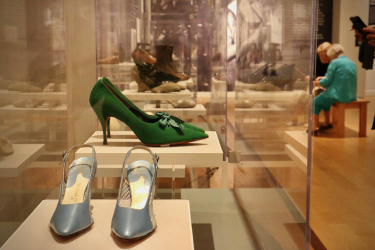 A pair of green high heels and silver high heels are in a glass display case. Museum-goers are visible in the background.