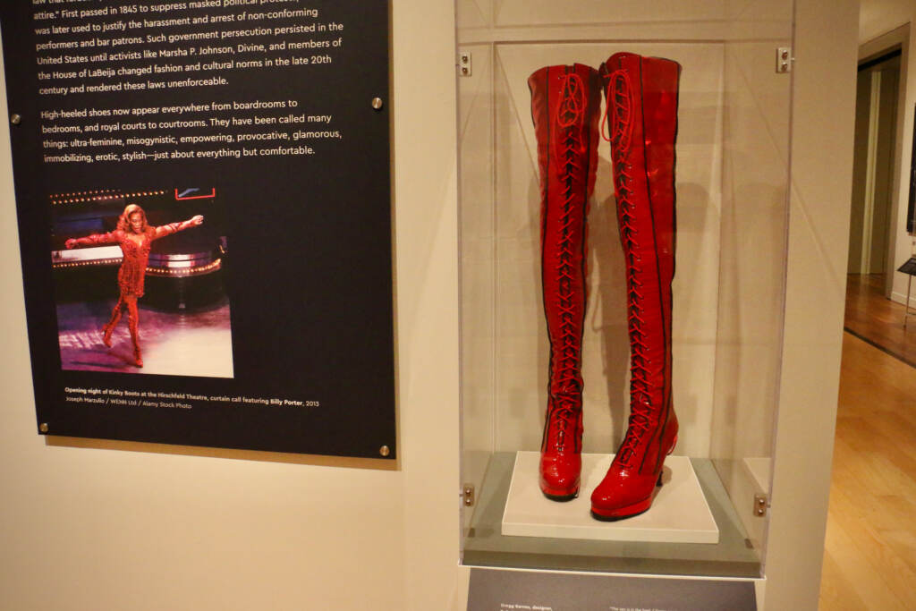 A glass display case holds a pair of leg-length red lace-up boots next to a sign that has words on it.
