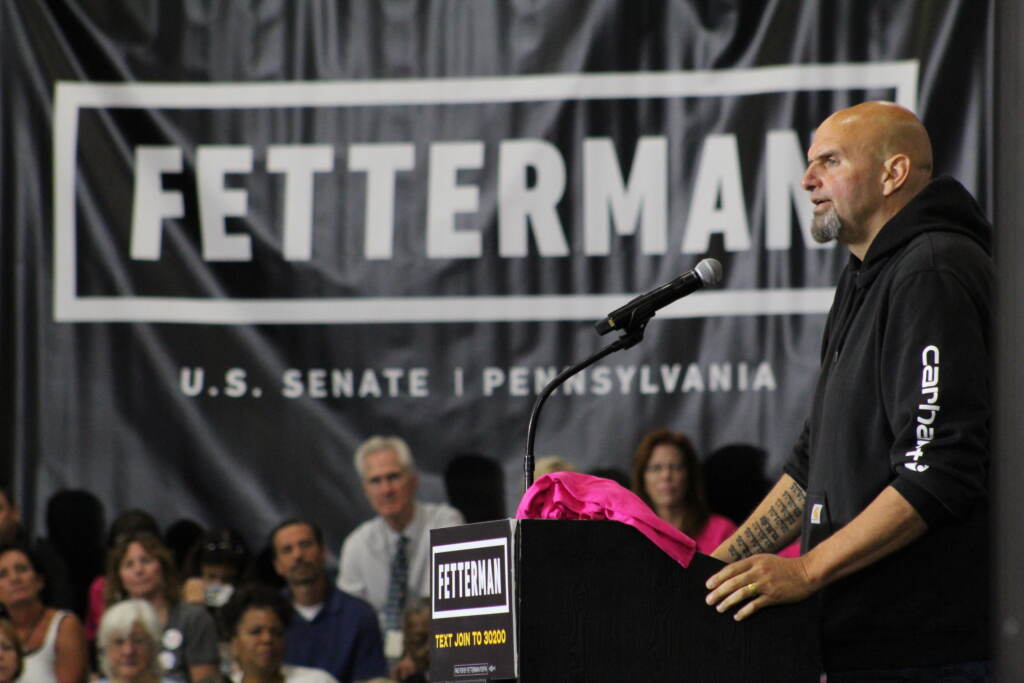 Fetterman-rally-5 Fetterman is visible in profile, speaking at a podium in front of a crowd. A huge Fetterman campaign sign hangs in the background.