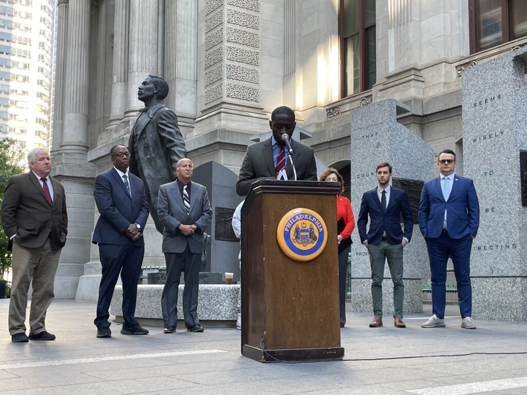 ThomasPresser Councilmember Thomas speaks at a podium outside of City Hall surrounded by other people.