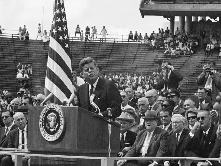A black-and-white photo of President Kennedy speaking in the midst of a large crowd.