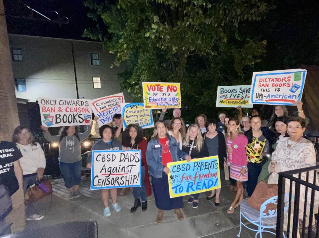 A group of people are gathered together at nighttime holding signs against censorship and banned books.