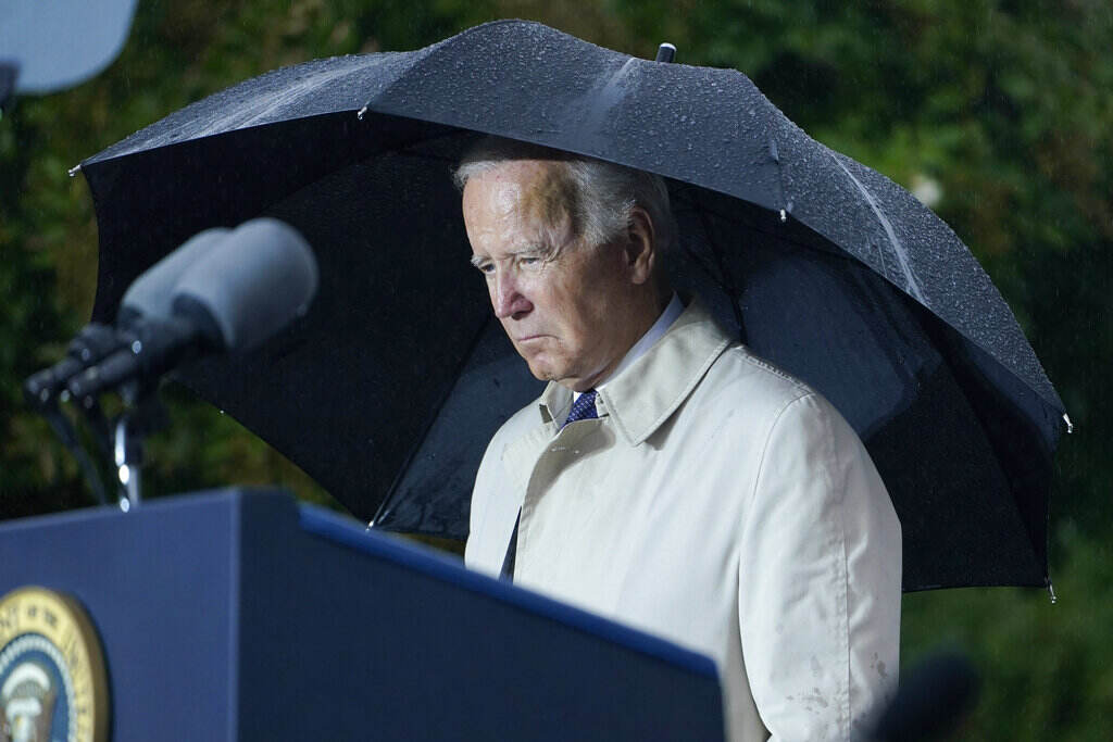 Joe Biden President Biden stands under a black umbrella at a podium, with a somber expression on his face.