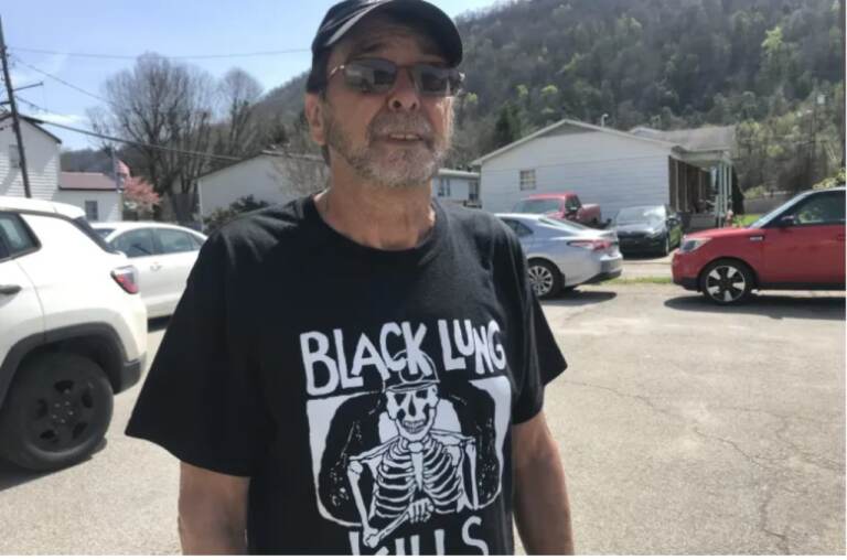 A man with a baseball hat and sunglasses faces the camera. He's wearing a shirt that reads, 